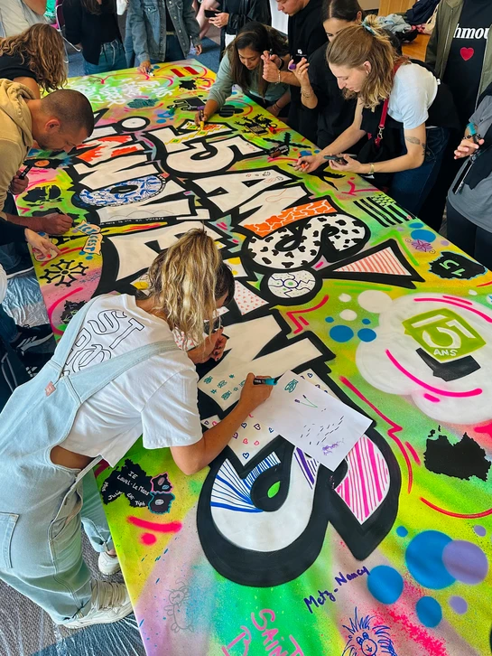 Groupe de professionnels en action lors d'une animation team building de graffiti : plusieurs personnes de tous âges, équipées de bombes de peinture, travaillent ensemble sur un grand mur blanc ou gris dans un espace intérieur type atelier. Certains sont en train de peindre, d'autres observent ou attendent leur tour, créant une atmosphère collaborative et ludique. L'ambiance doit être dynamique, professionnelle et conviviale, montrant l'aspect social et participatif de l'activité sans clichés banals.