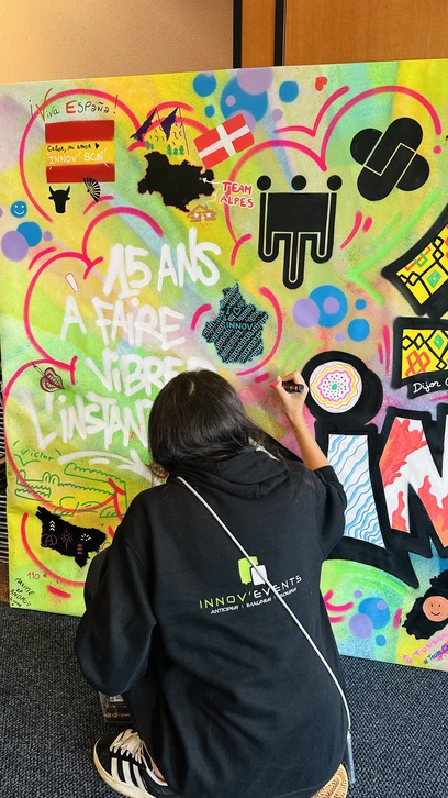 Photographie d'un groupe diversifié de professionnels en tenue décontractée collaborant activement à la création d'une fresque murale colorée. Certains membres de l'équipe tiennent des pinceaux et des bombes de peinture, appliquent des couleurs sur un mur blanc, tandis que d'autres observent avec engagement et participent au processus créatif. L'atmosphère est positive, collaborative et énergique, avec des sourires et une réelle concentration artistique. Le décor peut être un intérieur professionnel (mur d'un loft ou d'un espace d'événementiel) ou urbain. L'image doit capturer l'essence du live painting et de la fresque participative comme vecteur de cohésion d'équipe autour d'une création artistique commune.